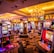 A vibrant casino interior filled with rows of colorful slot machines, each illuminated with bright lights and displays. The ceiling is adorned with elegant chandeliers and golden drapes, creating a luxurious atmosphere. The patterned carpet adds to the elaborate design of the space.