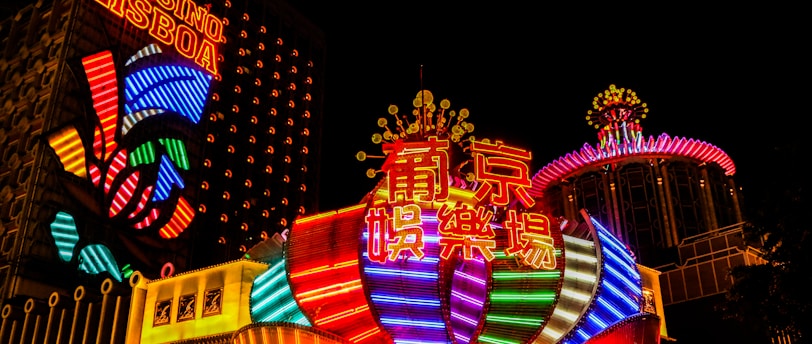 Bright neon lights and colorful signage adorn the exterior of Casino Lisboa, creating a vibrant and lively atmosphere. The façade features bold, multicolored designs with a mix of English and Chinese characters prominently displayed. The lights form intricate patterns and shapes, contributing to the energetic ambiance of the scene.