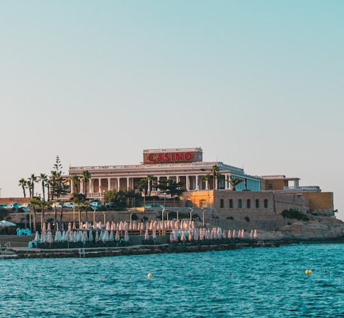 A seaside view of a large building labeled as a casino, situated on a rocky shore. The building is surrounded by palm trees and other smaller structures. The calm sea water reflects the sky, and there are several closed umbrellas lined along the waterfront.